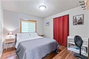 A bedroom with a bed, a bedside table, a lamp, a window with blinds, a red curtain, a wall-mounted picture, and a standing fan. - Stanley Stays - The Bonanza Cabin (Stanley)