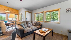 A cozy living room with a sofa, armchair, and coffee table, large windows with blinds, and a ceiling fan. - Stanley Stays - The Ponderosa Pine Cabin (Stanley)