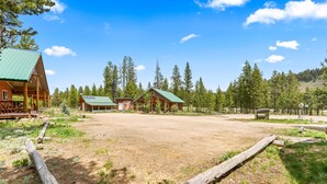 A rustic lodge with multiple cabins, a covered outdoor seating area, and a dirt ground surrounded by trees. - Stanley Stays - The Ponderosa Pine Cabin (Stanley)