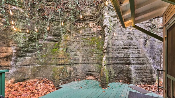 A rustic wooden deck with a green floor, surrounded by moss-covered rock walls and hanging light bulbs. - Cozy Eureka Springs Cottage, Walk to Dtwn! (Eureka Springs)