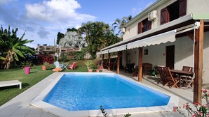 Una piscina con un porche techado, rodeada de plantas en macetas y mobiliario de exterior.
