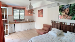 Un dormitorio con cama, una silla, un ventanal y una zona de cocina con armarios y un lavabo.