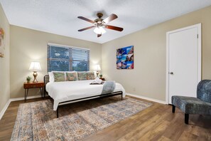 Ein Schlafzimmer mit Bett, Deckenventilator, Fenster mit Jalousien, Teppich, Nachttisch mit Lampe und einer Tür.