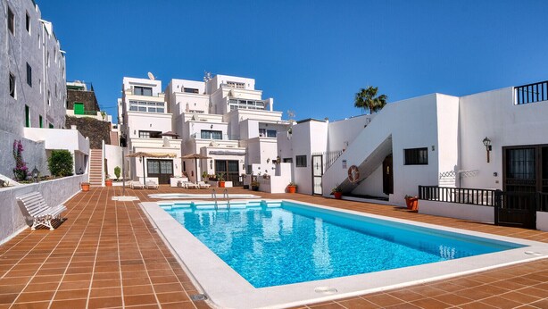 A swimming pool with a tiled deck, white buildings, and a clear blue sky. - Sea Waves - 500M from the beach, Pool and Terrace By LVH (Puerto del Carmen)