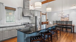 A modern kitchen with a large island, stainless steel appliances, wooden bar stools, and a dining area with a wooden table and chairs. - Bon Temps | 5 Bedroom w/Private Pool (Rosemary Beach)