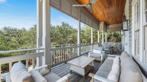 A covered porch with white wicker furniture, a wooden table, and a view of trees. - Bon Temps | 5 Bedroom w/Private Pool (Rosemary Beach)