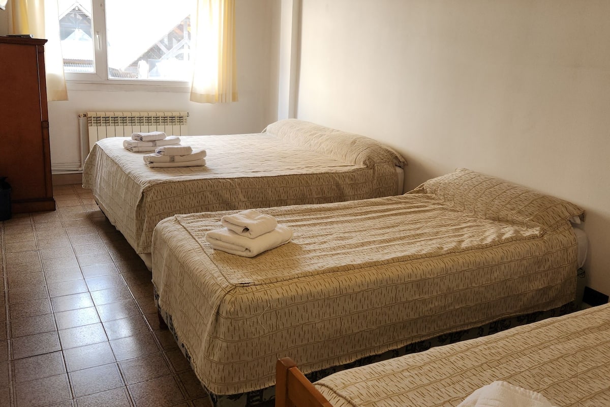 A room with two beds, a wooden ceiling, a window with curtains, and a radiator.