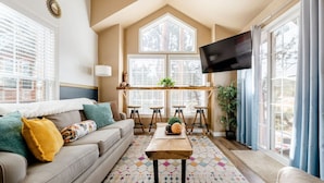 A cozy living room with a sofa, coffee table, and bar stools. There is a TV mounted on the wall and a window with blinds. - Cozy 1BR Chalet | Pikes Peak, Grill & Relaxing Patio (Woodland Park)