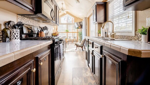 A kitchen with dark wood cabinets, a central island with a sink, and a view of the dining area through large windows. - Cozy 1BR Chalet | Pikes Peak, Grill & Relaxing Patio (Woodland Park)