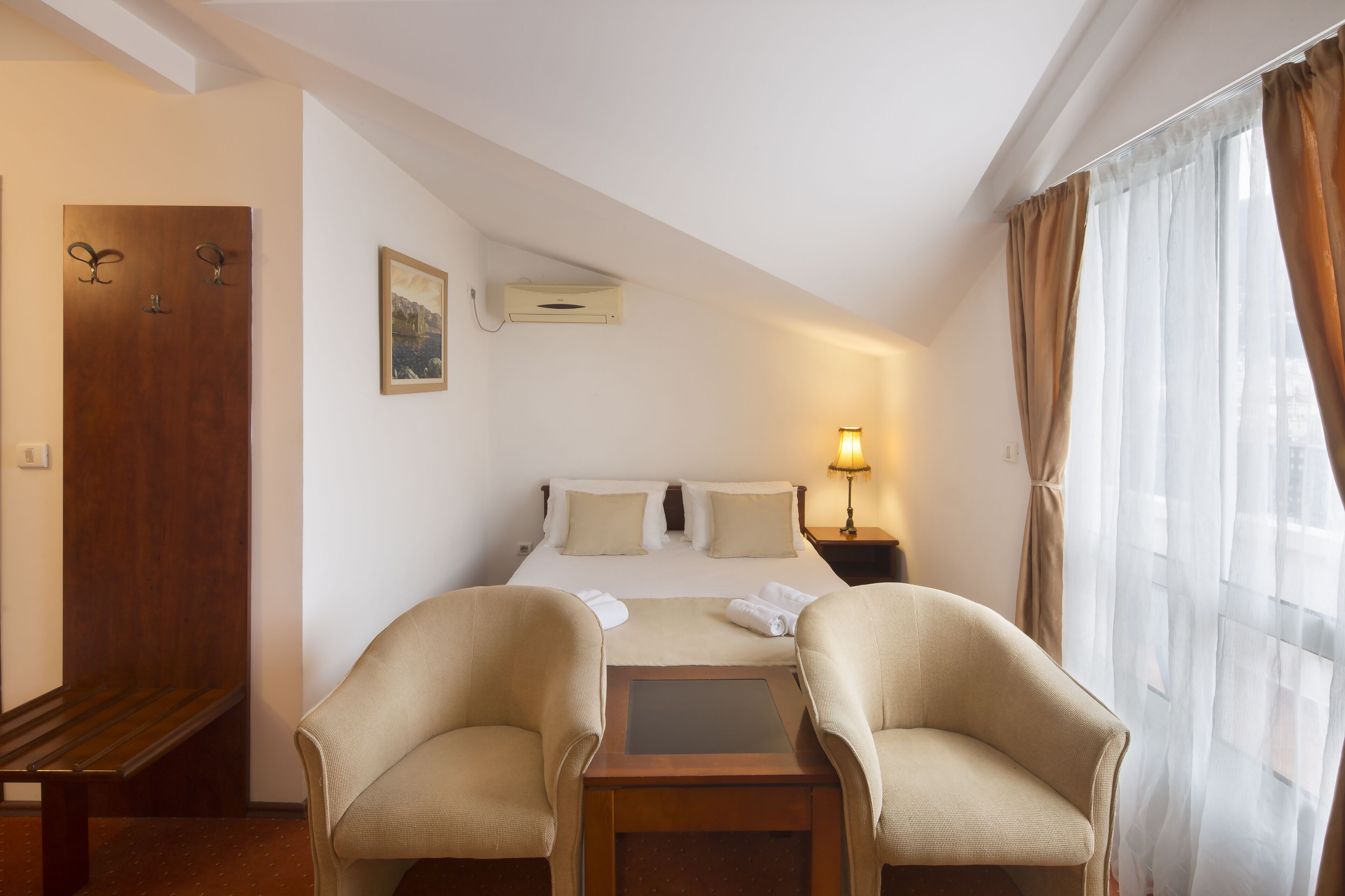 A hotel room with a bed, two armchairs, a small table, a nightstand with a lamp, a wooden door, and a window with curtains.