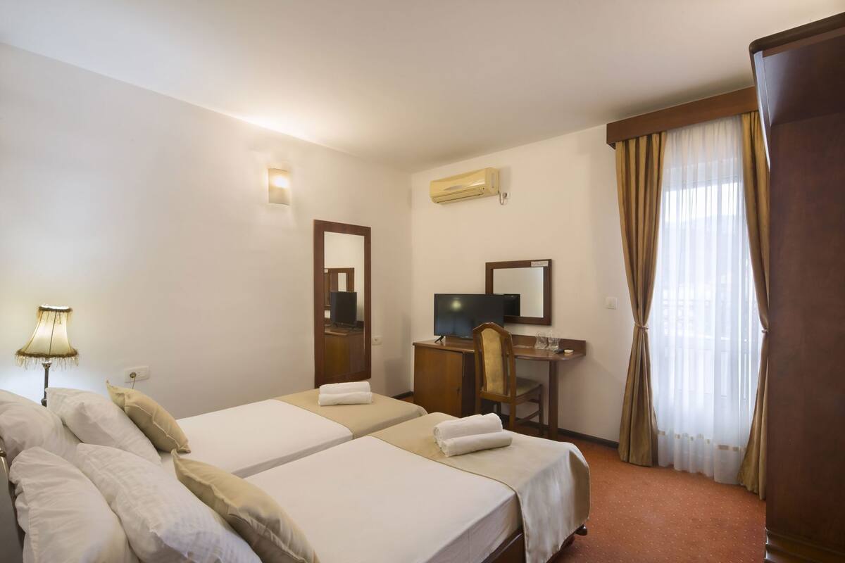 A hotel room with two beds, a desk with a computer, a mirror, an air conditioner, and a window with curtains.