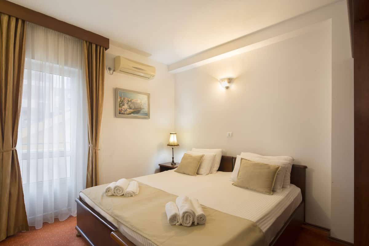 A hotel room with a bed, curtains, a lamp, a painting, and a wall-mounted air conditioner.