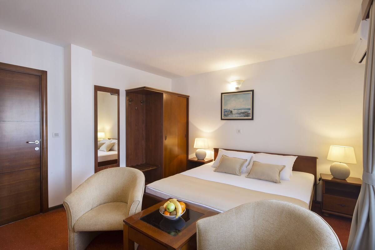 A hotel room with a bed, two chairs, a small table with fruit, a painting on the wall, and a door.
