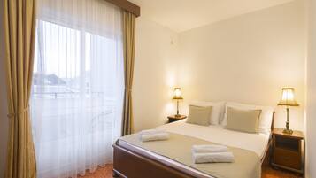 A hotel room with a large bed, two bedside tables with lamps, a window with sheer curtains, and a view of buildings outside.