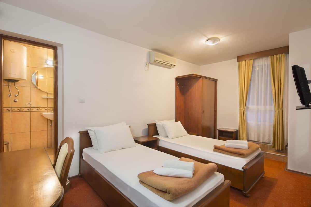 A hotel room with two beds, a wooden wardrobe, a desk, a chair, a television, and a bathroom with a mirror and sink.
