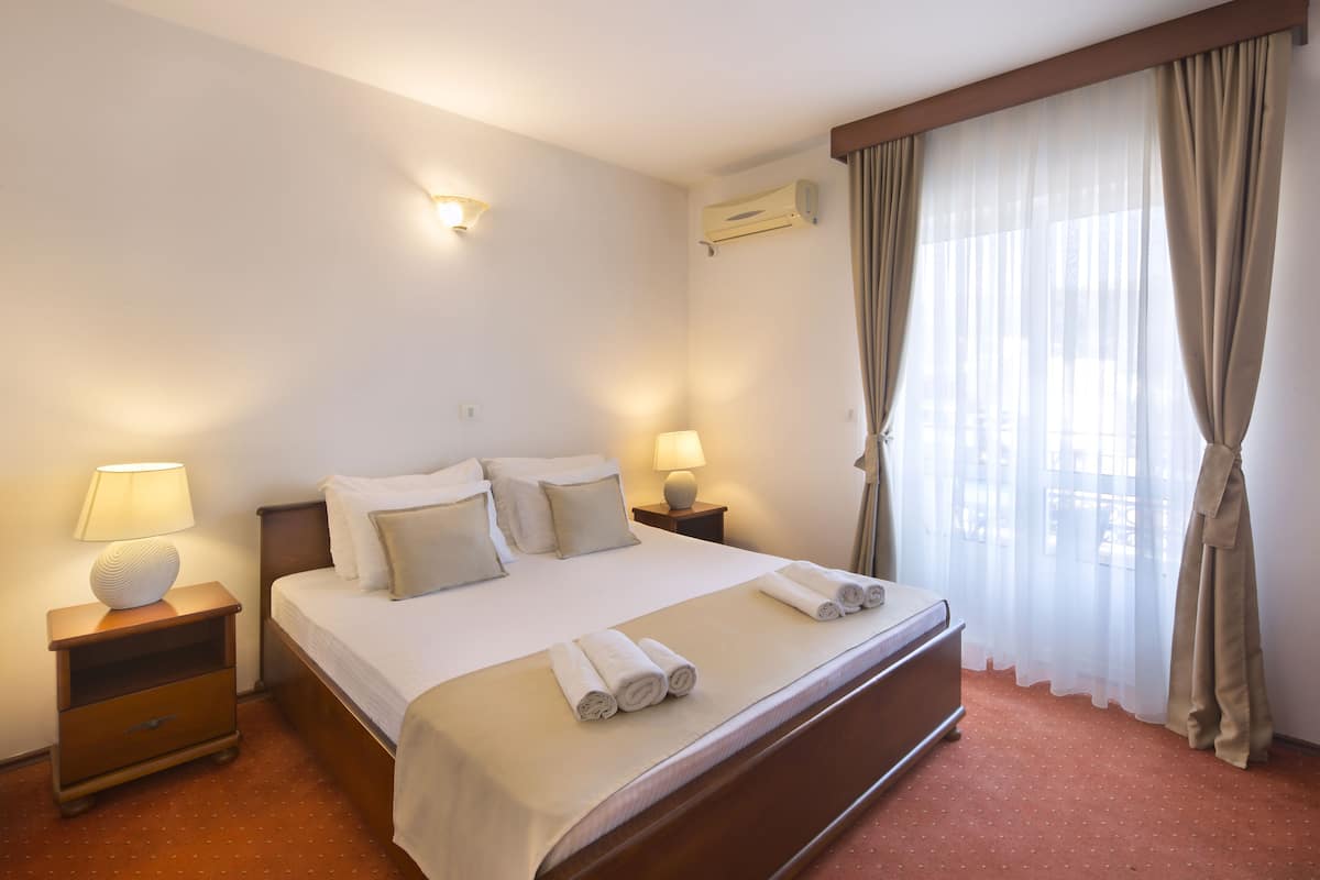 A hotel room with a bed, two bedside tables with lamps, a window with sheer curtains, and a wall-mounted air conditioner.