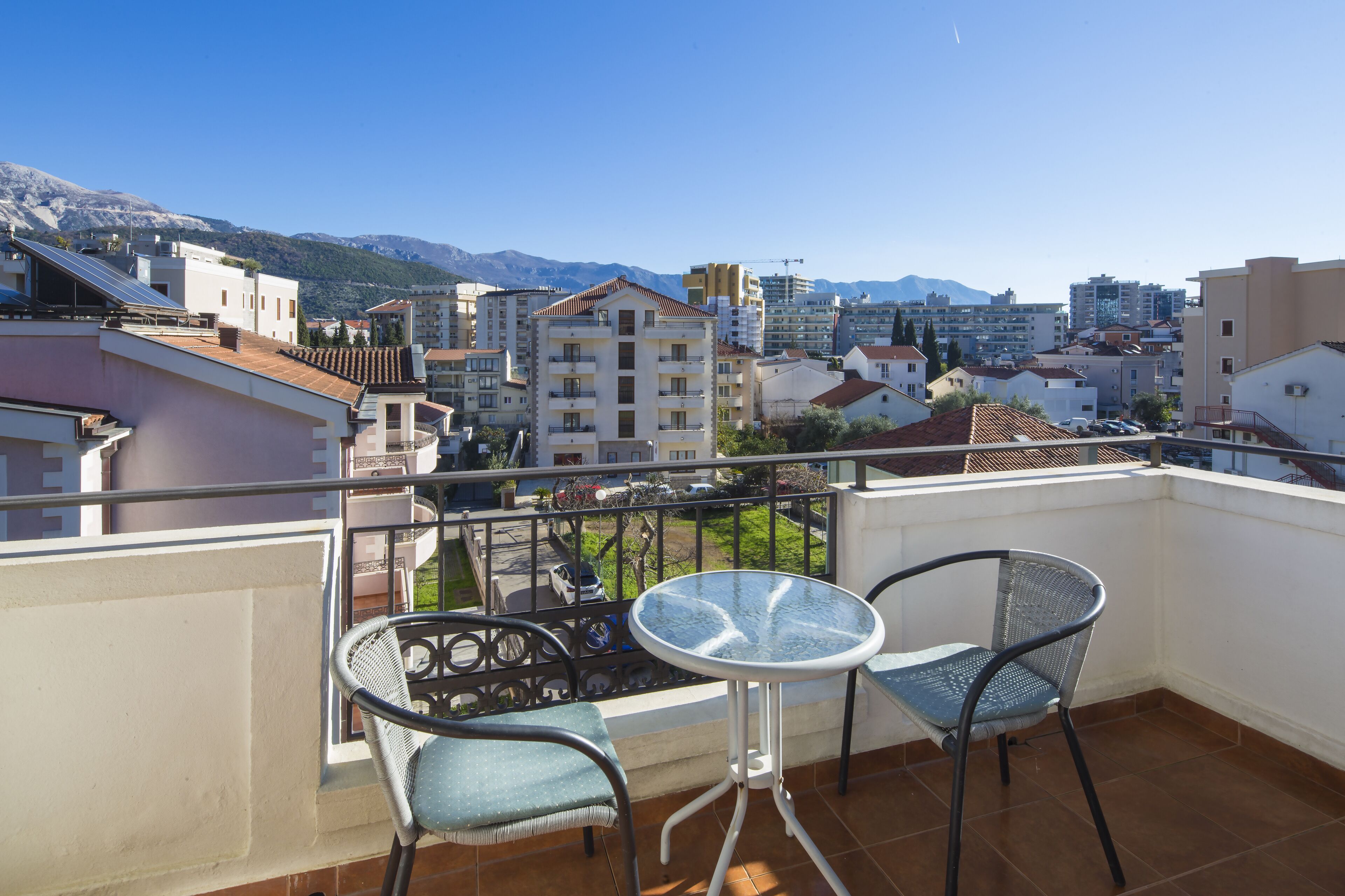 A balcony with two chairs and a table, overlooking a cityscape with buildings and mountains.
