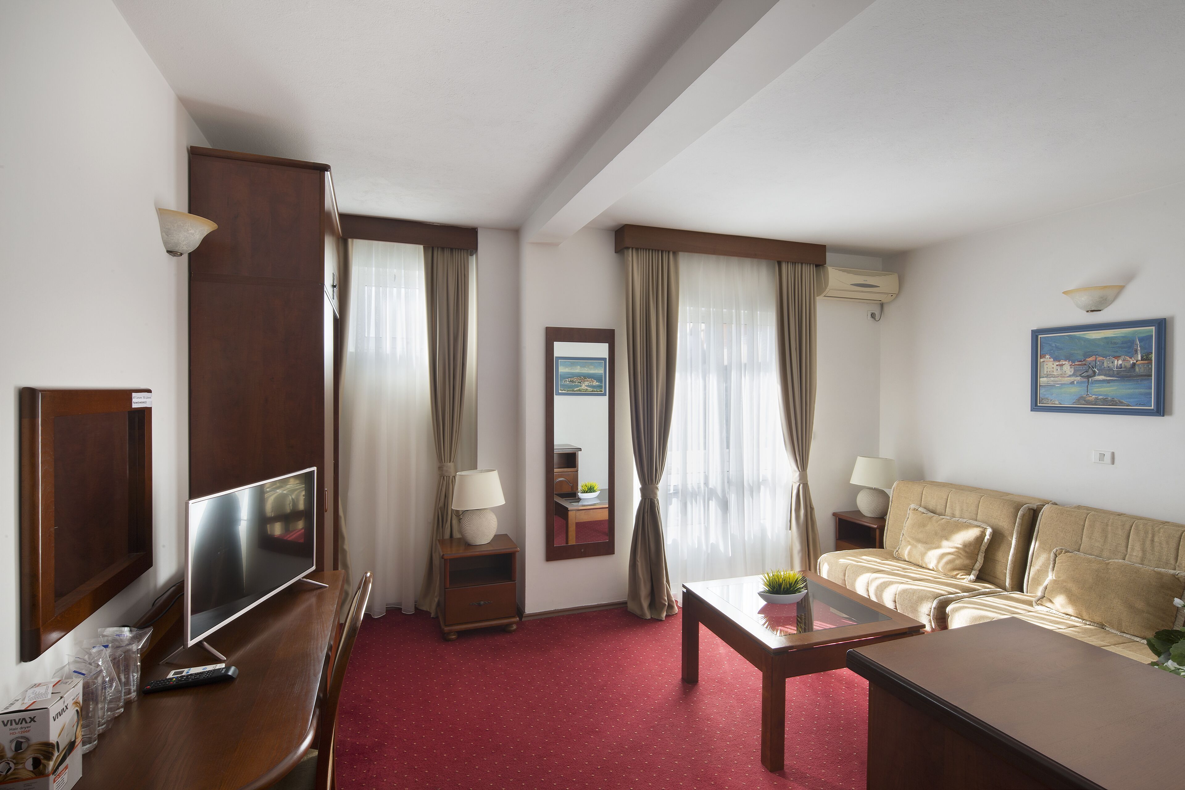 A hotel room with a TV, wooden furniture, a red carpet, a sofa, and a painting on the wall.