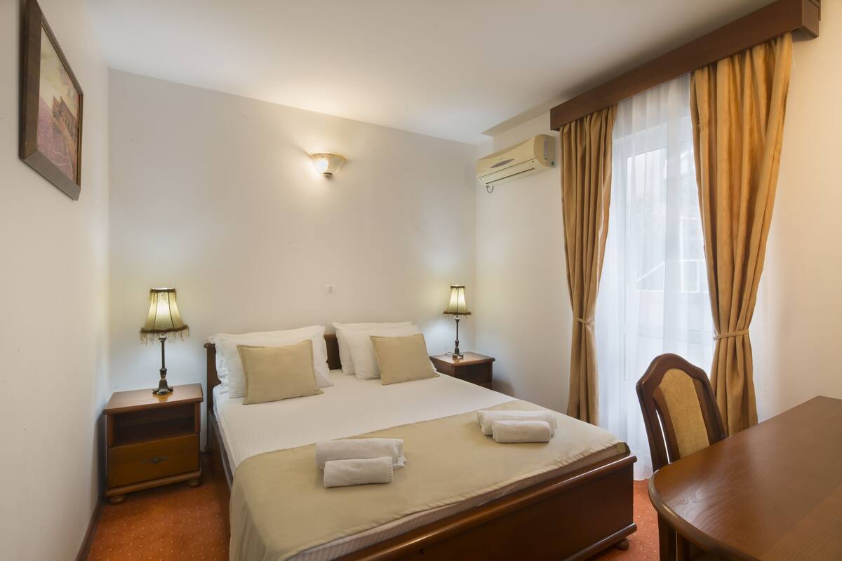 A hotel room with a bed, two bedside tables with lamps, a chair, a table, and a window with curtains.