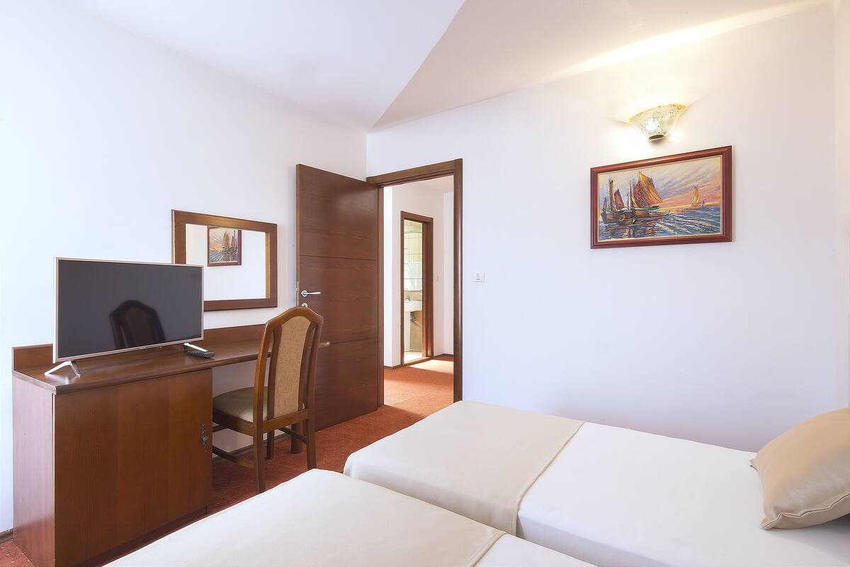A hotel room with a bed, a desk with a computer, a chair, a painting, and a door leading to another room.
