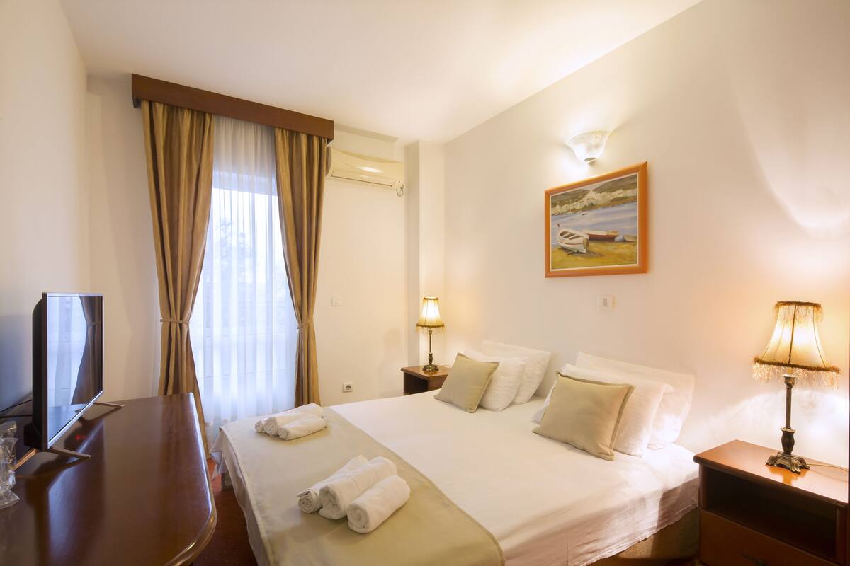 A hotel room with a bed, a TV, a desk, and a painting on the wall.