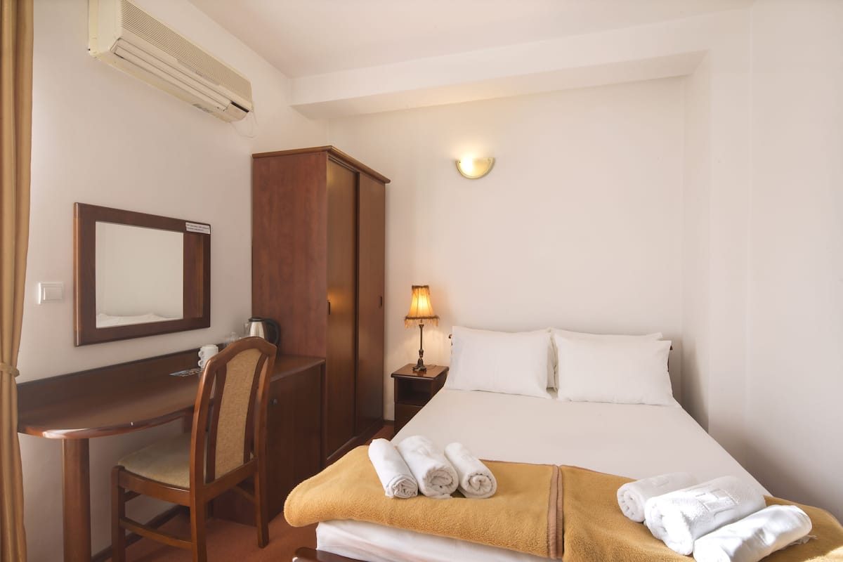 A hotel room with a bed, a desk, a chair, a nightstand with a lamp, and a mirror.