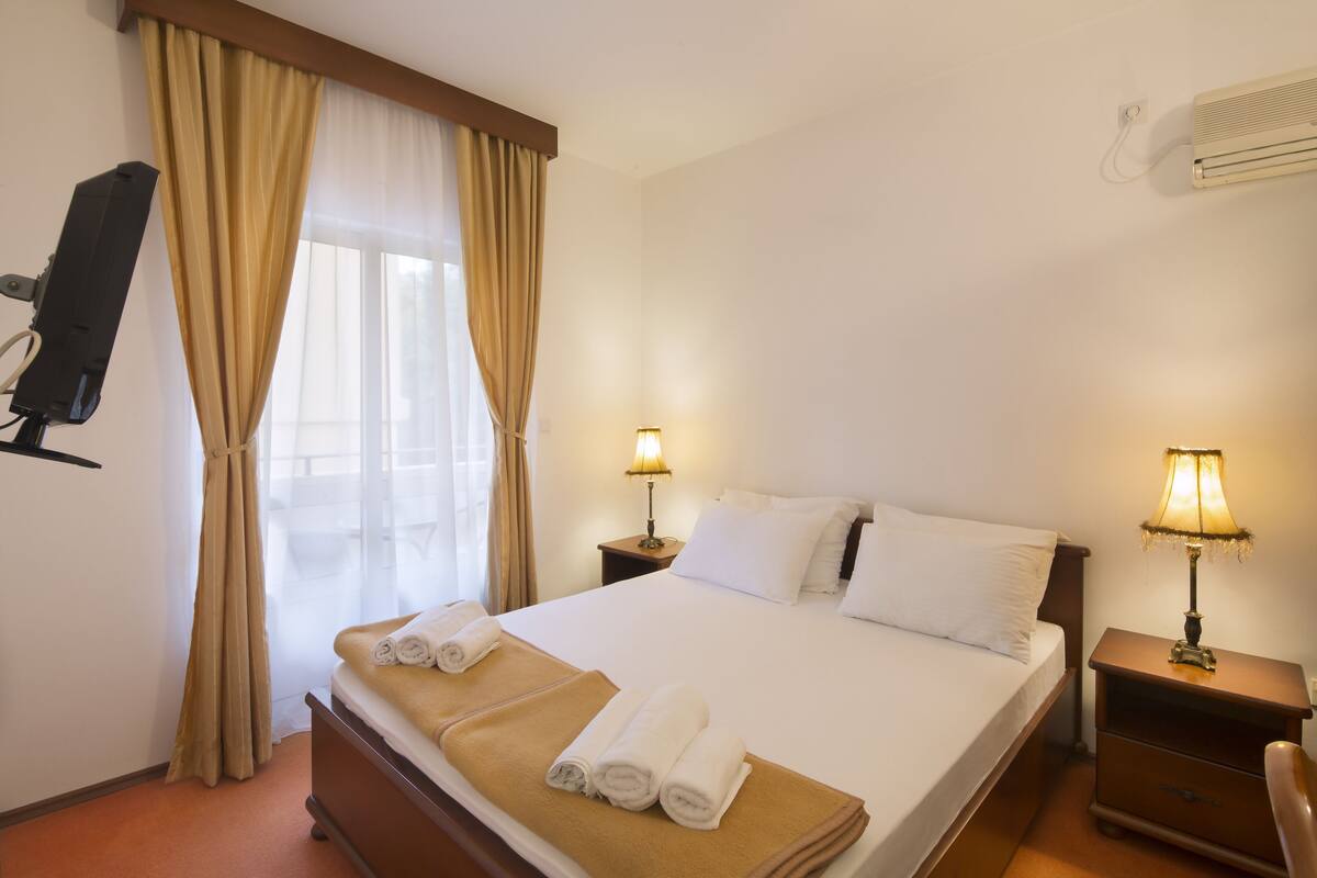 A hotel room with a bed, two bedside lamps, a TV, and a window with curtains.