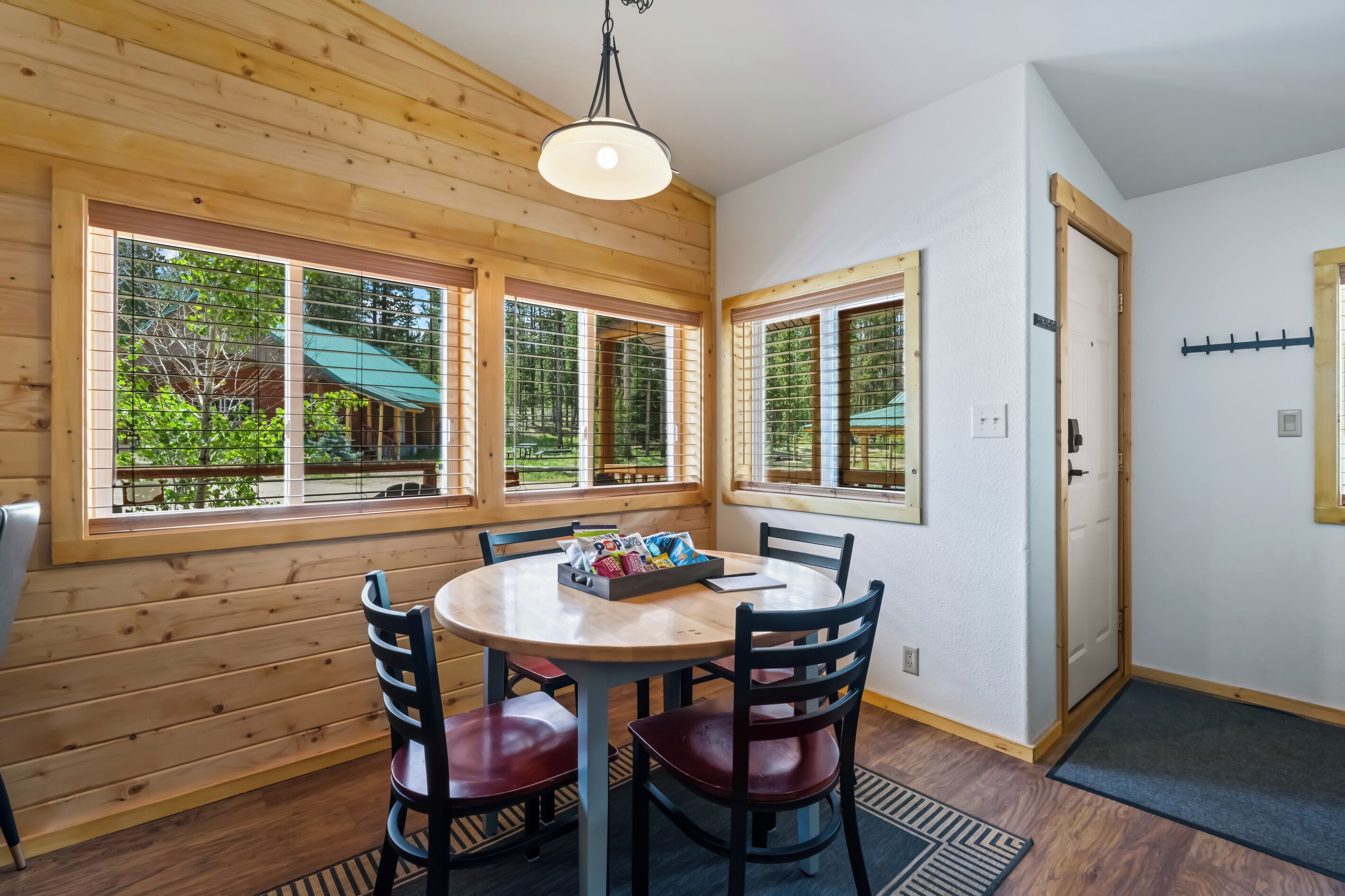 A wooden-paneled room with a round table and chairs, large windows with a view of greenery, and a door with a coat rack.