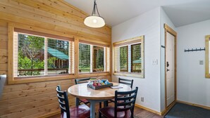 Habitación con paneles de madera, una mesa redonda y sillas, ventanas amplias con vistas a la vegetación, y una puerta con perchero.