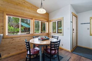 A wooden-paneled room with a round table and chairs, large windows with a view of greenery, and a door with a coat rack. - Stanley Stays - The Valley Creek Cabin (Stanley)