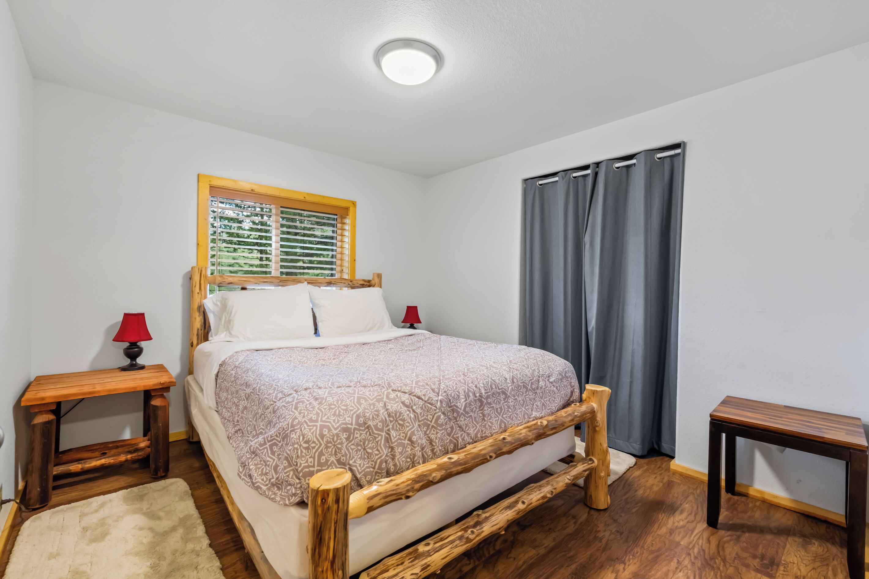 Una camera da letto con un letto con struttura in legno, una finestra con tende, un comodino con una lampada e uno sgabello di legno.