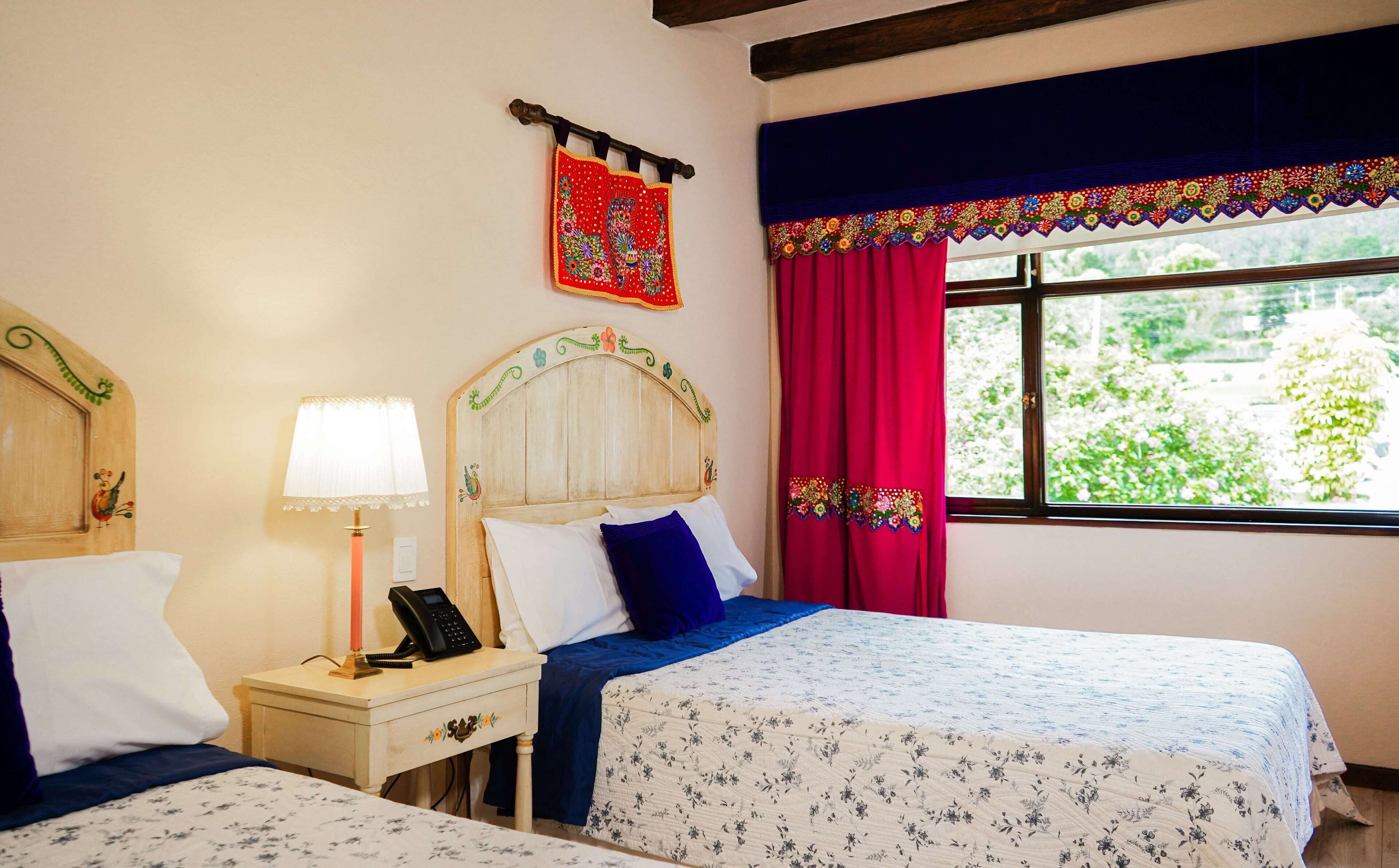 Una habitación con dos camas, cabecera de madera con adornos florales, mesita de noche con lámpara y teléfono, y ventana con vista a la vegetación.