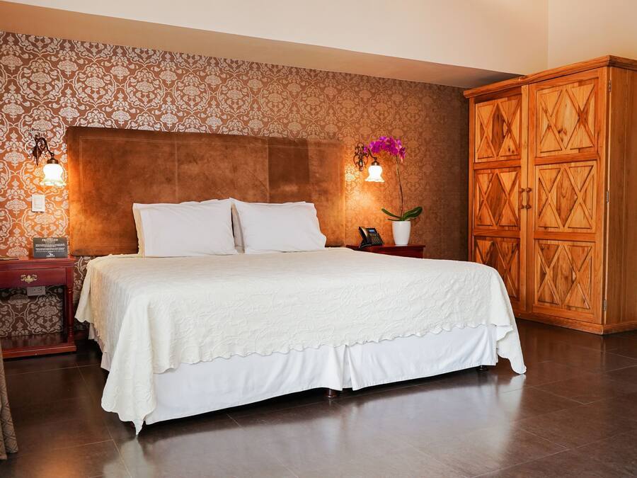 A neatly made bed with a white bedspread, a wooden headboard, a nightstand with a lamp, a telephone, and a decorative vase with an orchid.