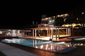 Una piscina amb zona coberta i seients de relax, envoltada d'un pati ben il·luminat i un edifici al fons.