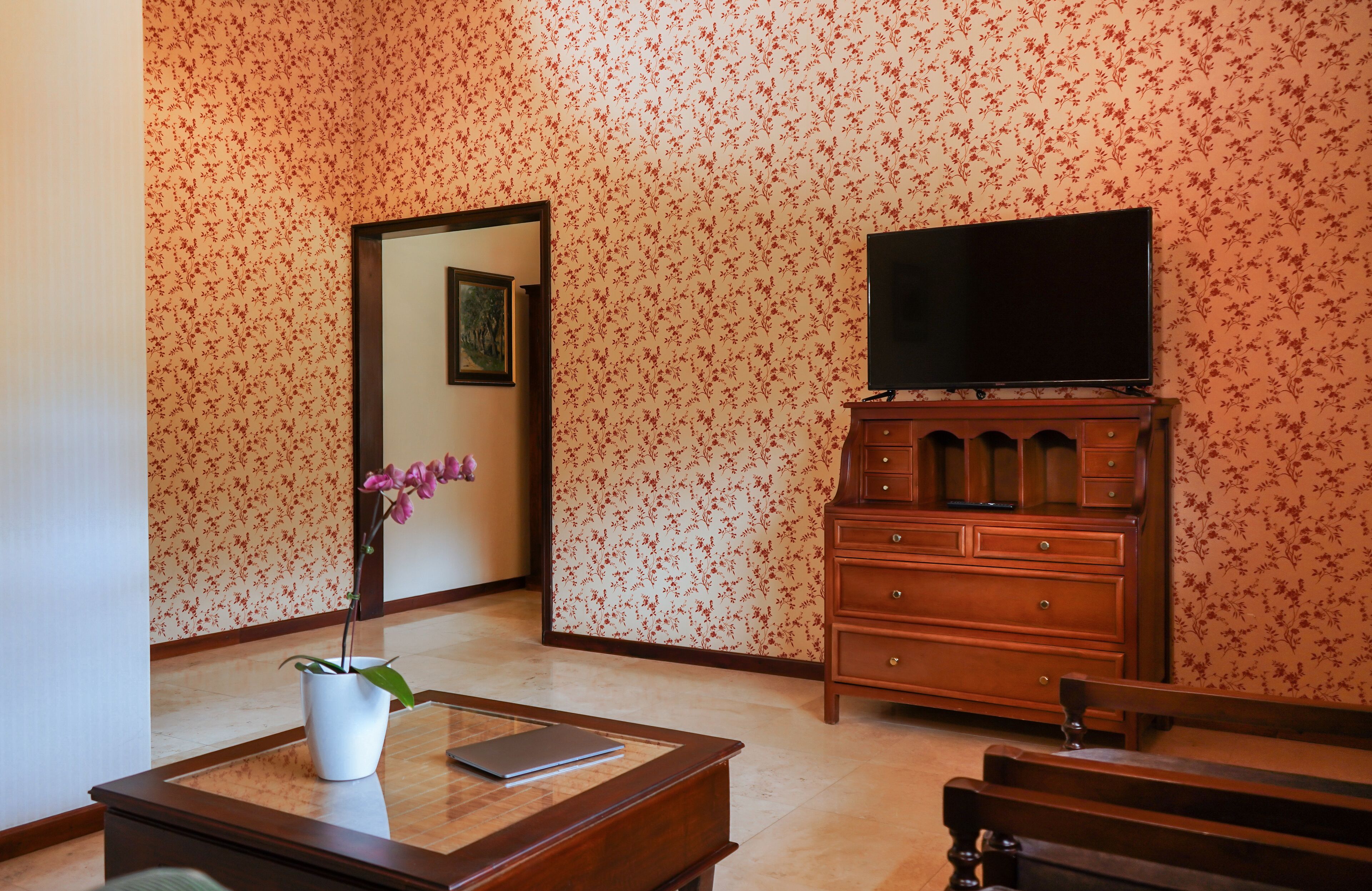 A room with floral wallpaper, a wooden dresser, a television, a desk with a laptop, a bench, and a potted orchid.