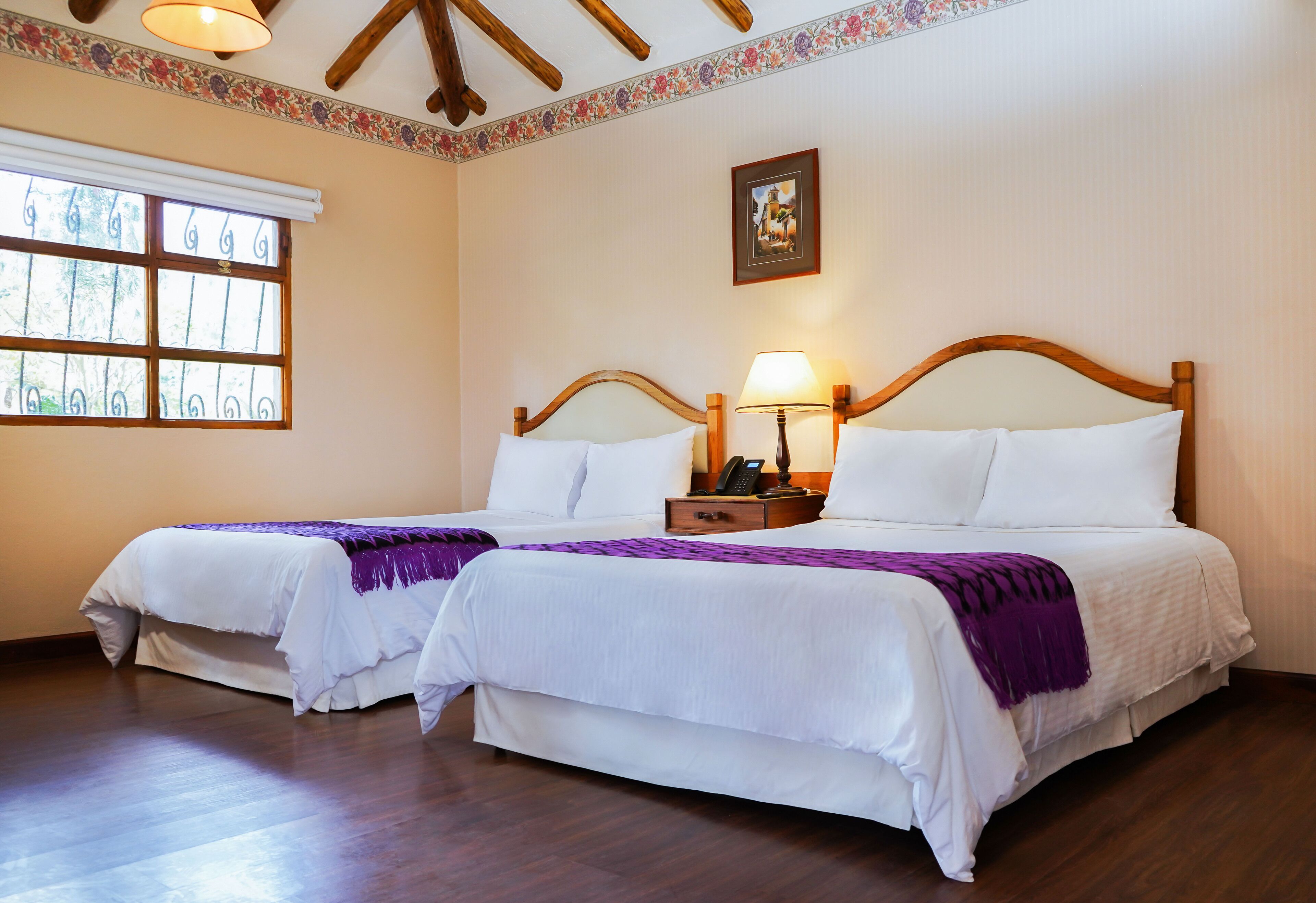 Habitación de hotel con dos camas, cabeceros de madera, una mesita de noche con lámpara, una ventana con rejas y un borde floral en el techo.