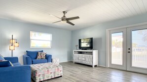 A living room with a blue sofa, a patterned ottoman, a white entertainment unit, and a ceiling fan. - Upper-Level 2BR | Pool, Private Deck & 2-Min Walk to Beach and Flagler Avenue (New Smyrna Beach)