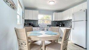 A modern kitchen with white cabinets, a central island, and a dining area with four chairs and blue placemats. - Upper-Level 2BR | Pool, Private Deck & 2-Min Walk to Beach and Flagler Avenue (New Smyrna Beach)
