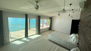 Camera d'albergo con due letti, un balcone vista mare, ventilatori a soffitto e lampade a sospensione.