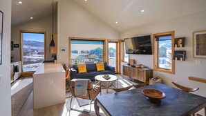 A spacious living area with a sofa, coffee table, and dining table. A kitchen area with a refrigerator and cabinets is visible. Large windows offer views of a snowy landscape.