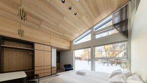 A modern bedroom with a large bed, a desk, and a chair. There are multiple windows providing natural light and a view of the outside.