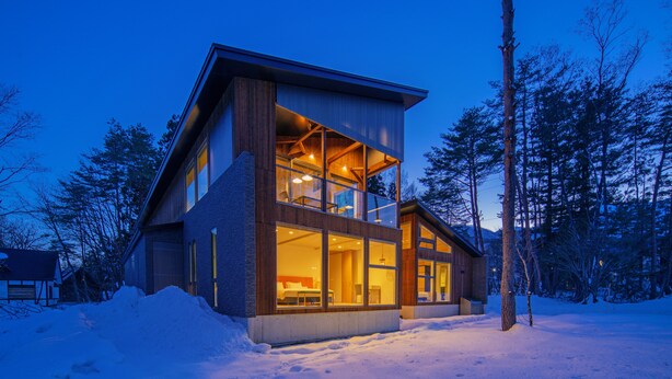 A modern two-story cabin with large windows and a wooden exterior, surrounded by snow and trees.