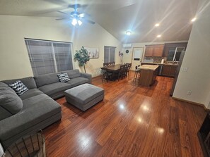 A spacious living room with a gray sectional sofa, a dining area with a wooden table and chairs, and a kitchen area with wooden cabinets and a countertop.