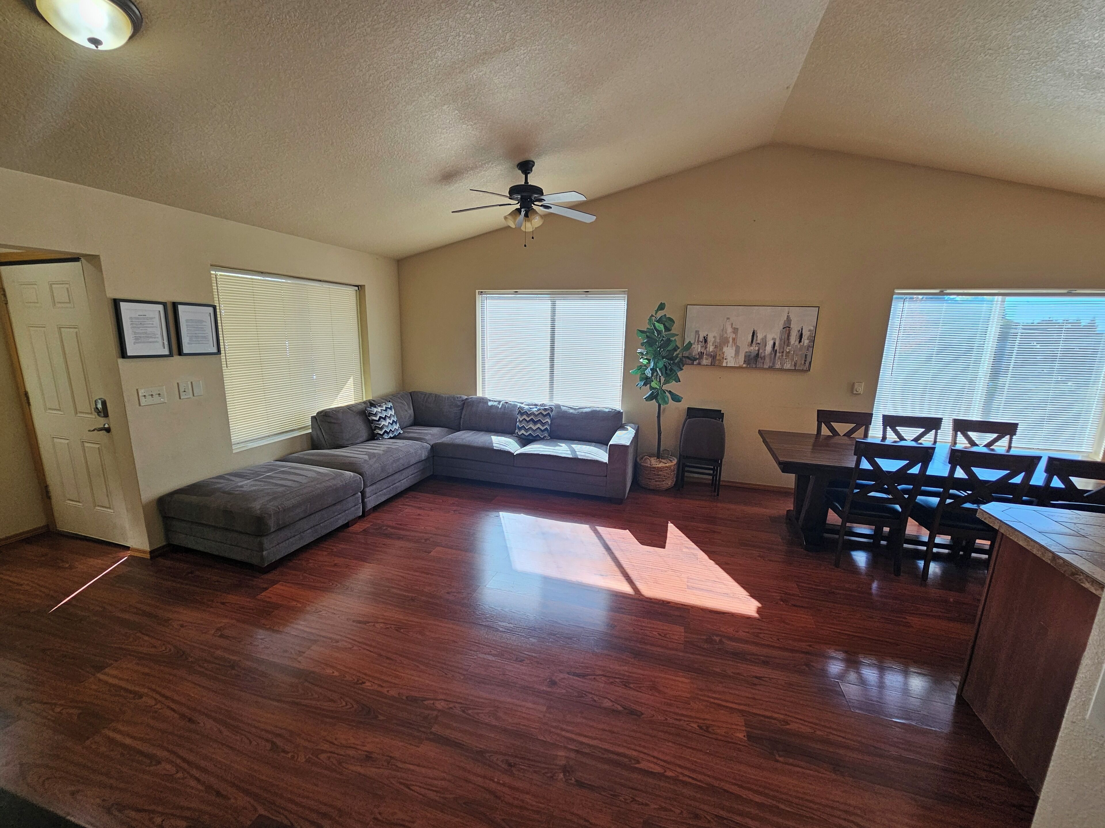 A living room with a sectional sofa, a dining table with chairs, a ceiling fan, and a cityscape painting.