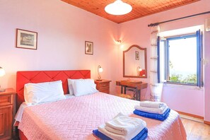 A bedroom with a red headboard, a bed with white and pink bedding, a wooden nightstand, a desk with a chair, a mirror, and a window with curtains. - Old Stone House in Nisaki (Corfu)
