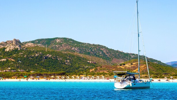 A sailboat on a clear blue sea with a mountainous backdrop and a beach with umbrellas. - Piazzetta Bilo Porto 50m From The Beach (Loiri Porto San Paolo)