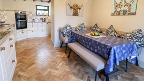 A modern kitchen with built-in appliances, a dining area with a bench and table, and a wall with a painting of donkeys. - 3 Penllan Cottages (Holywell)