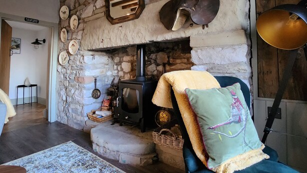 A rustic room with a stone fireplace, a wooden ceiling, and a cozy seating area with a chair and cushions. - 3 Penllan Cottages (Holywell)