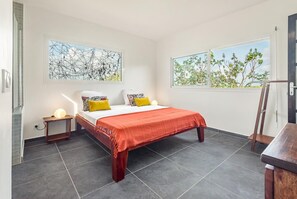 Un dormitorio moderno con una cama grande, dos mesitas de noche con lámparas, una silla de madera y dos ventanas amplias con vistas a árboles.