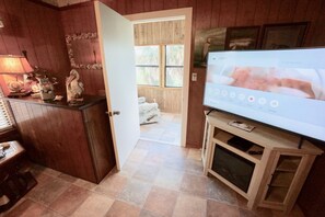 A room with a wooden floor, a television on a wooden stand, a door leading to another room, and a small table with a lamp and decorative items.